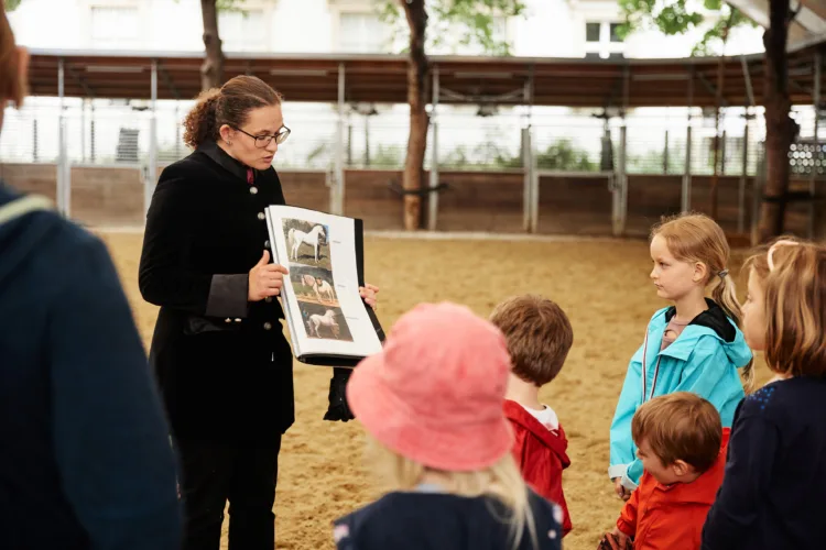 eine Gruppe von Kindern steht in einer offenen Reitbahn, die von Bäumen und einer Führanlage gesäumt ist. Sie blicken zu einer jungen Frau im Gehrock, die anhand von Bildern etwas erklärt. Auf den Bildern sind Pferde dargestellt.