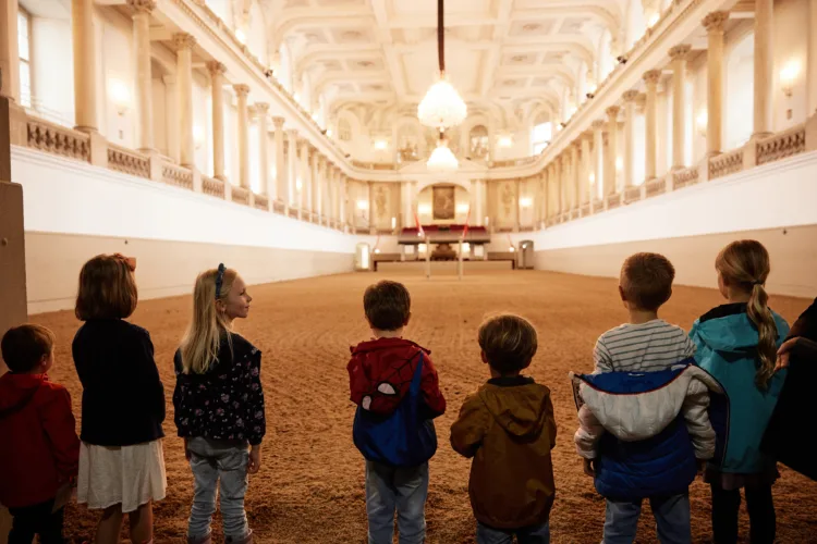 eine Gruppe von Kindern ist von hinten zu sehen. Sie blicken in eine barocke Reithalle, die hell erleuchtet ist. Sie stehen am Eingang in die Halle am Reitboden.