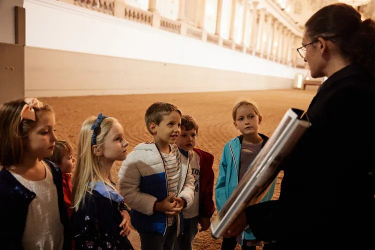 eine Gruppe von Kindern steht in einer hellen Reithalle und blickt interessiert zu einer jungen Frau, die ihnen einen Ordner mit Bildern zeigt und etwas zu erklären scheint.