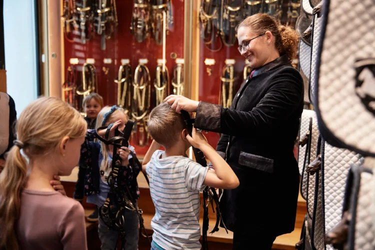 mehrere Kinder sind in einer Sattelkammer zu sehen. Manche von ihnen probieren mit einer jungen Frau spielerisch Zaumzeuge aus.