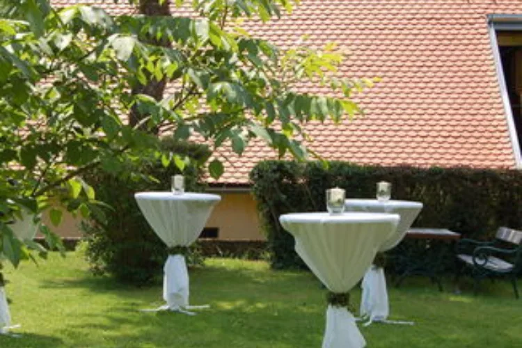 Green area with green trees in the background, festively decorated bar tables with white covers, building roof in the background