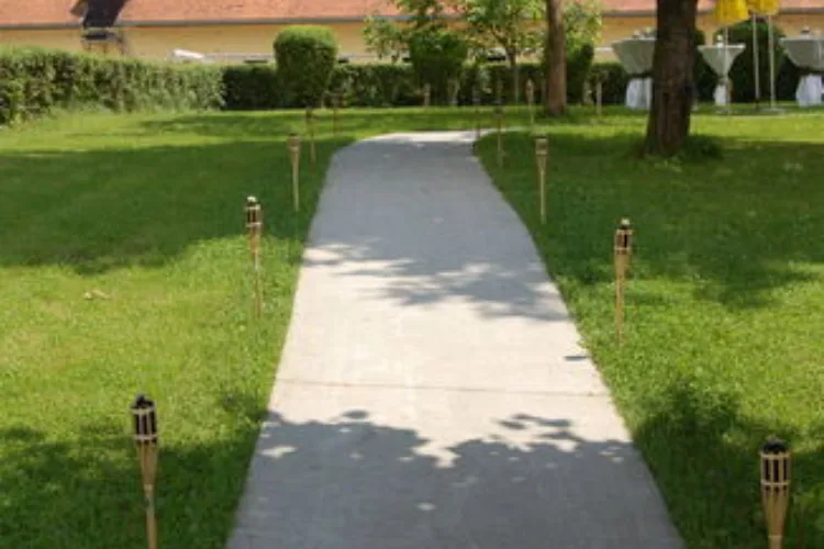 Path with torches by the wayside, sunshine, shade from trees, green meadow, a yellow building in the background