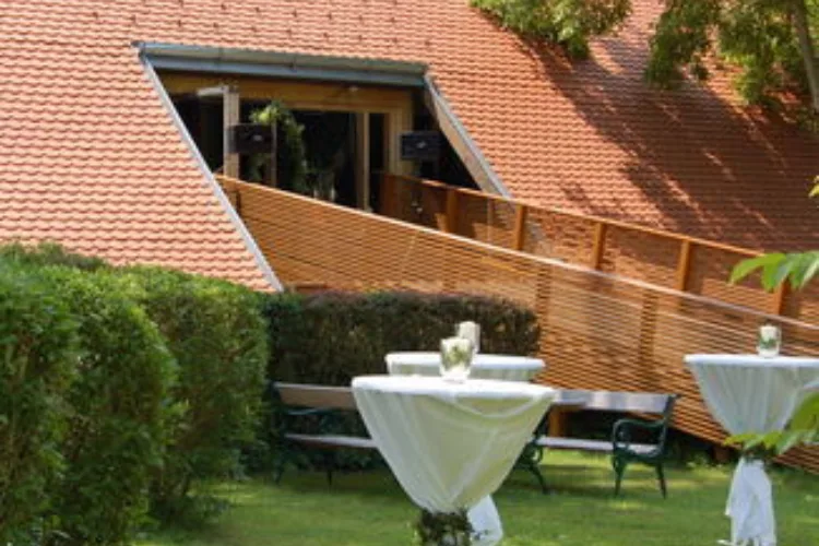 Green area with bar tables with white, festive covers, behind them the roof of a building